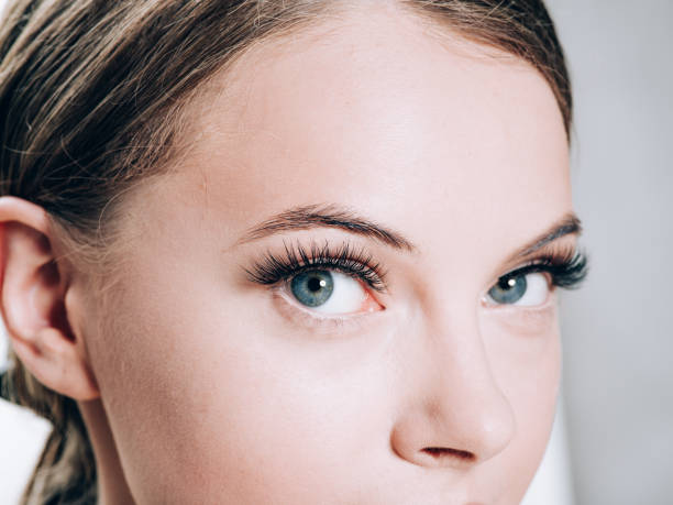 Close-up portrait of a woman with striking blue eyes and well-defined eyelashes, showing detailed facial features against a soft gray background