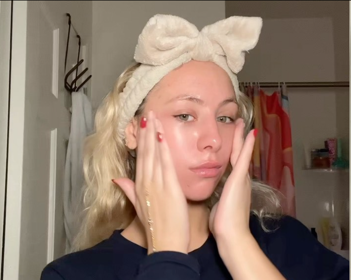 Woman wearing a headband applying skincare product to her face in a bathroom setting.