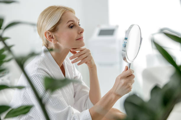 A woman in a white bathrobe looking at her reflection in a handheld mirror, sitting in a serene, modern room with plants